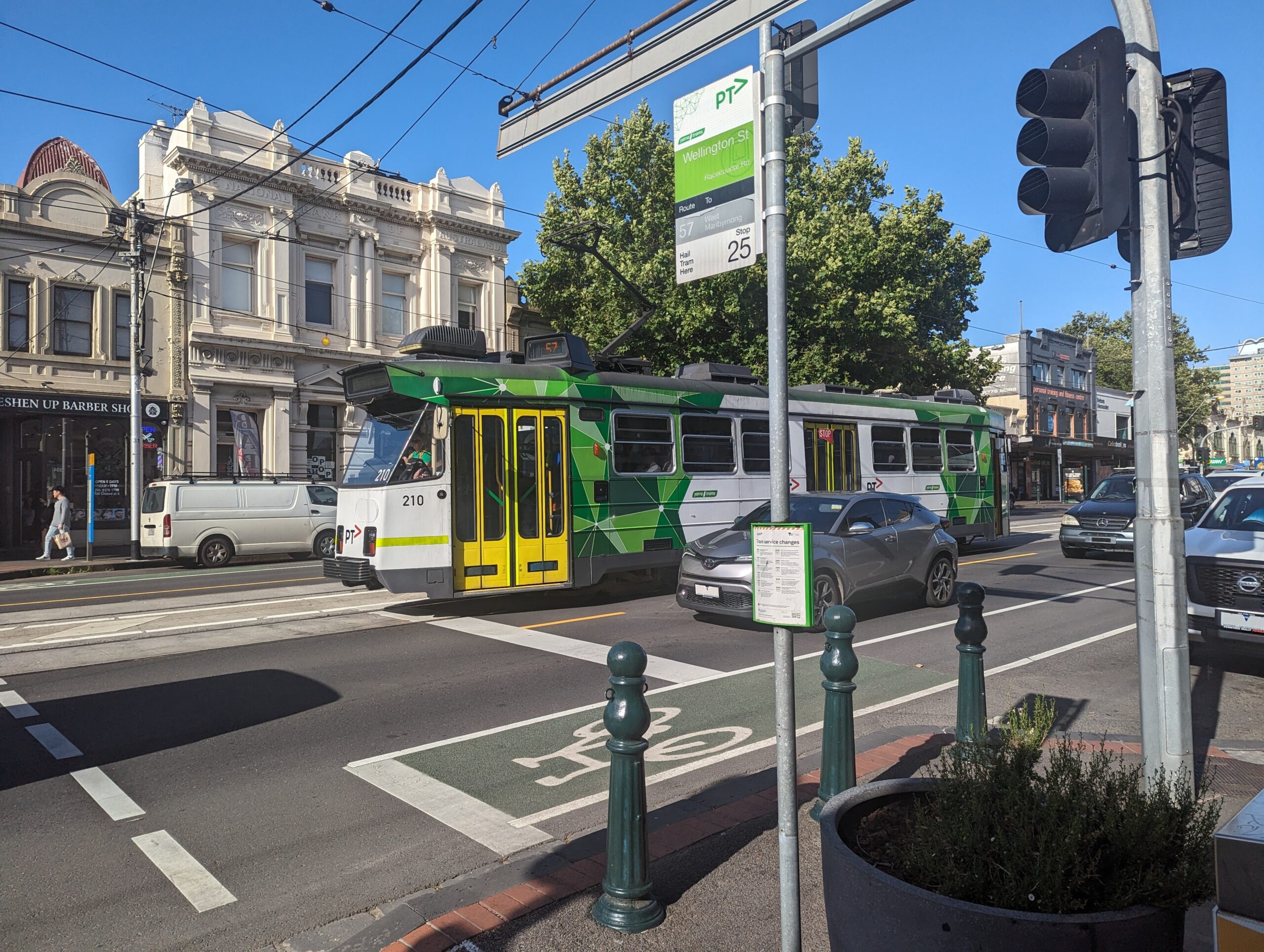 A high-floor tram sits in the middle of a street, among cars. A pole with a sign denotes the tram stop; there is no platform or other infrastructure.