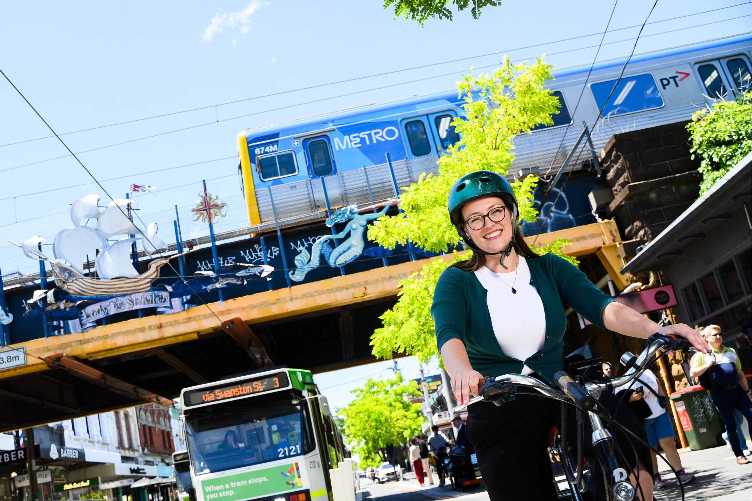 Katherine Copsey MLC sitting on a bike in a protected bike lane, with a tram at a stop behind her, while a train crosses a bridge overhead