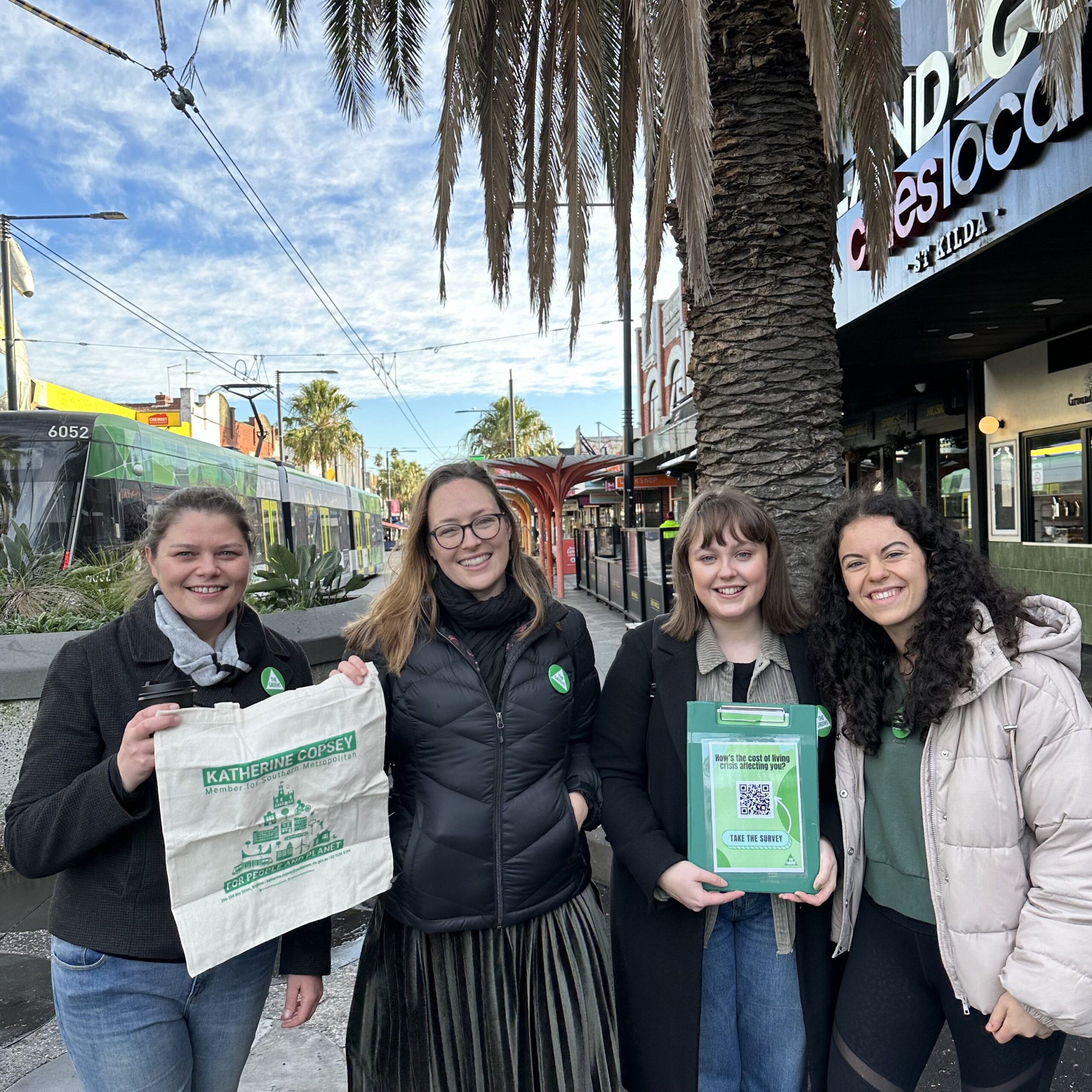 Katherine Copsey MP stands with volunteers at a Cost of Living stall. A volunteer on the left is holding a Katherine Copsey tote bag, while one on the right is holding a green clipboard with a QR code for the cost of living survey